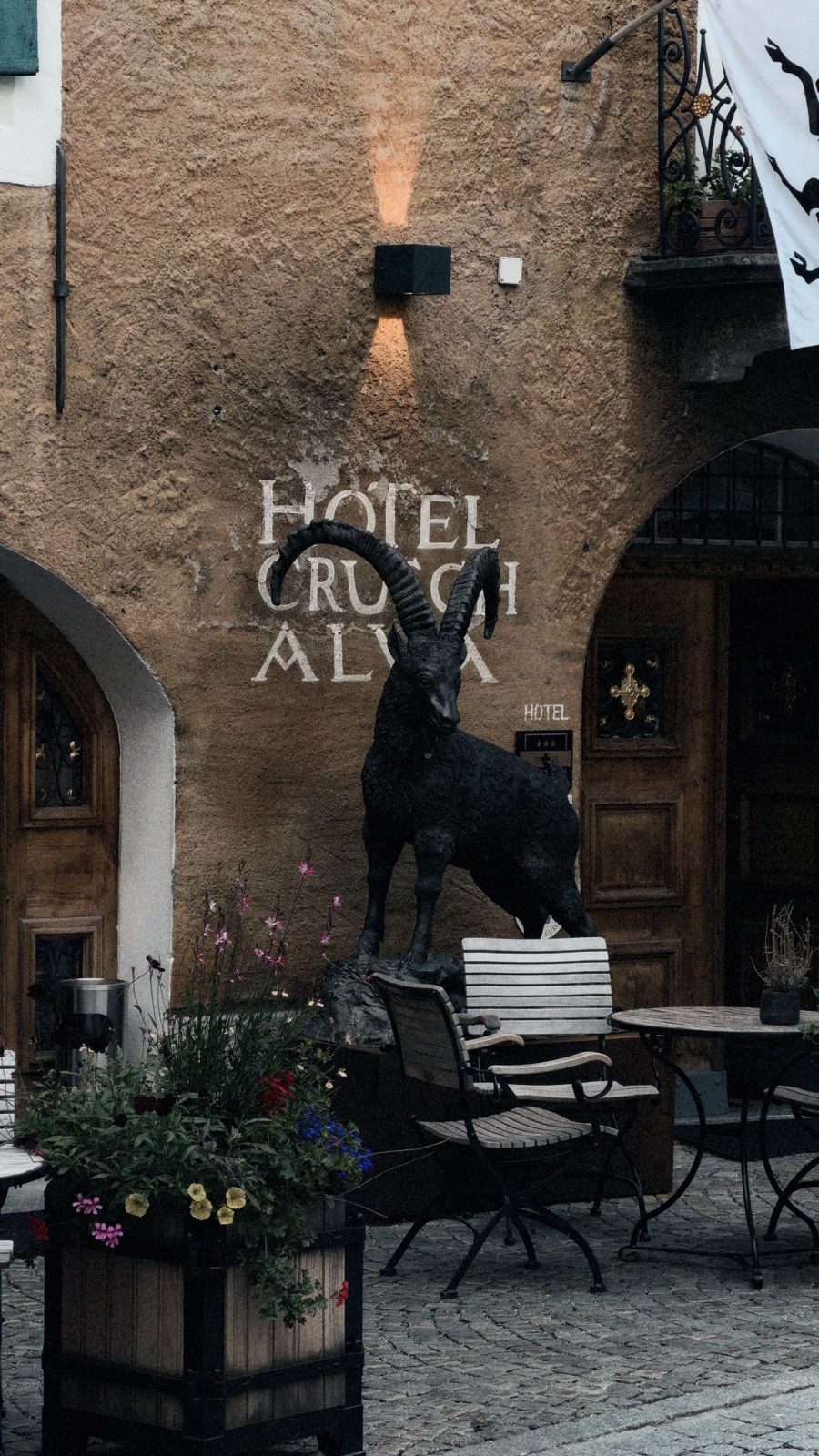 Steinernes Haus mit dem Schriftzug Hotel Crusch Alva, einer Steinbockstatue und Gartentischen